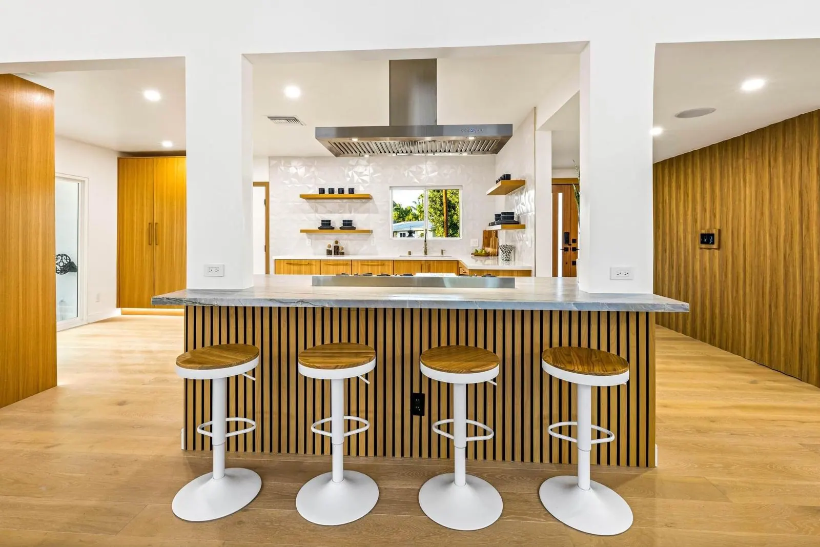 Wood kitchen with a slatted breakfast bar, white stools, a stone countertop and a stainless range hood